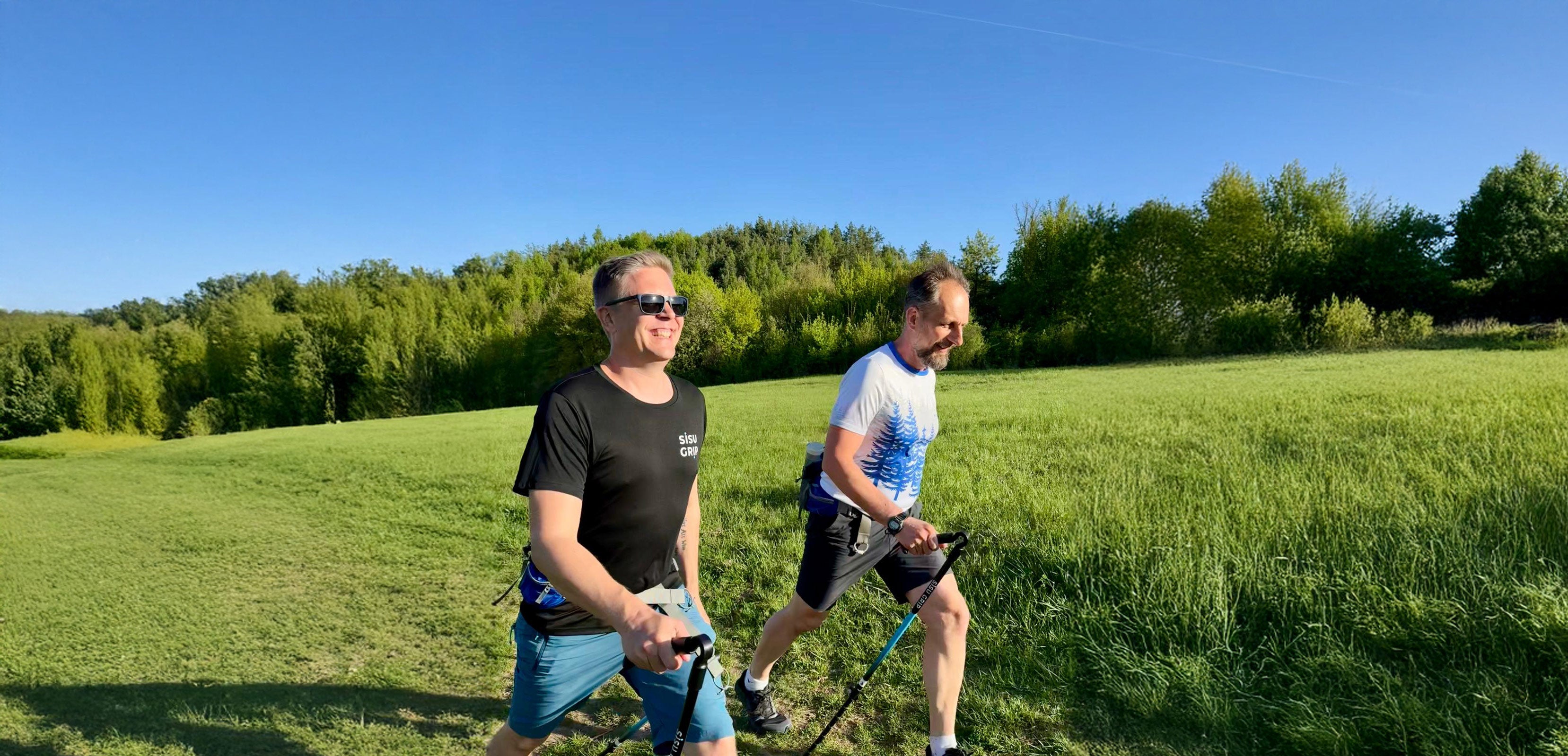 Kaksi miestä kävelee Sisu Grip sauvoilla niityn reunassa